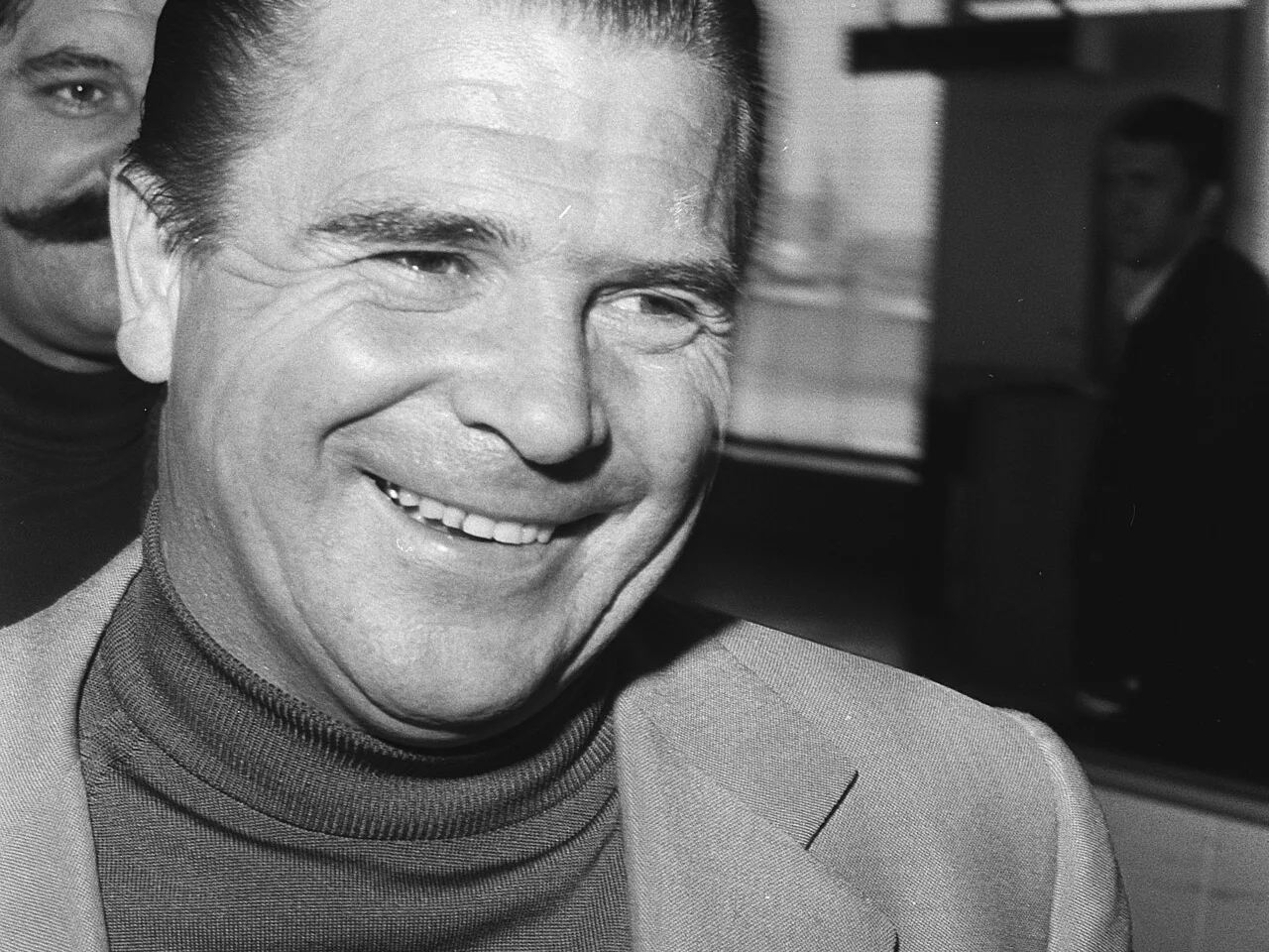 Ferenc Puskás ler i polotröja och kavaj vid ankomsten till Schiphol 1971, då som tränare för Panathinaikos